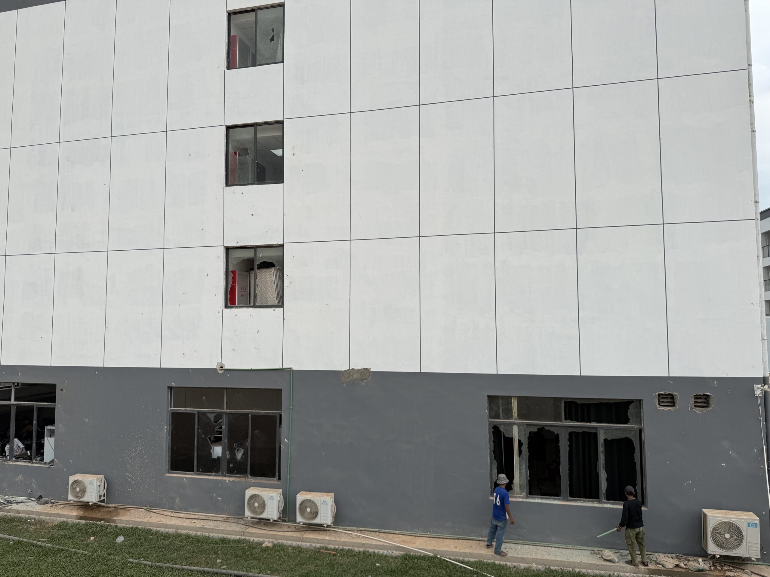 Two men look through broken windows on the side of a compound called Prek Khsach on February 12, 2026, after foreign and local construction workers allegedly broke them in protest of unpaid wages in Cambodia's Koh Kong province. (Mech Dara/Mekong Independent/Creative Commons)