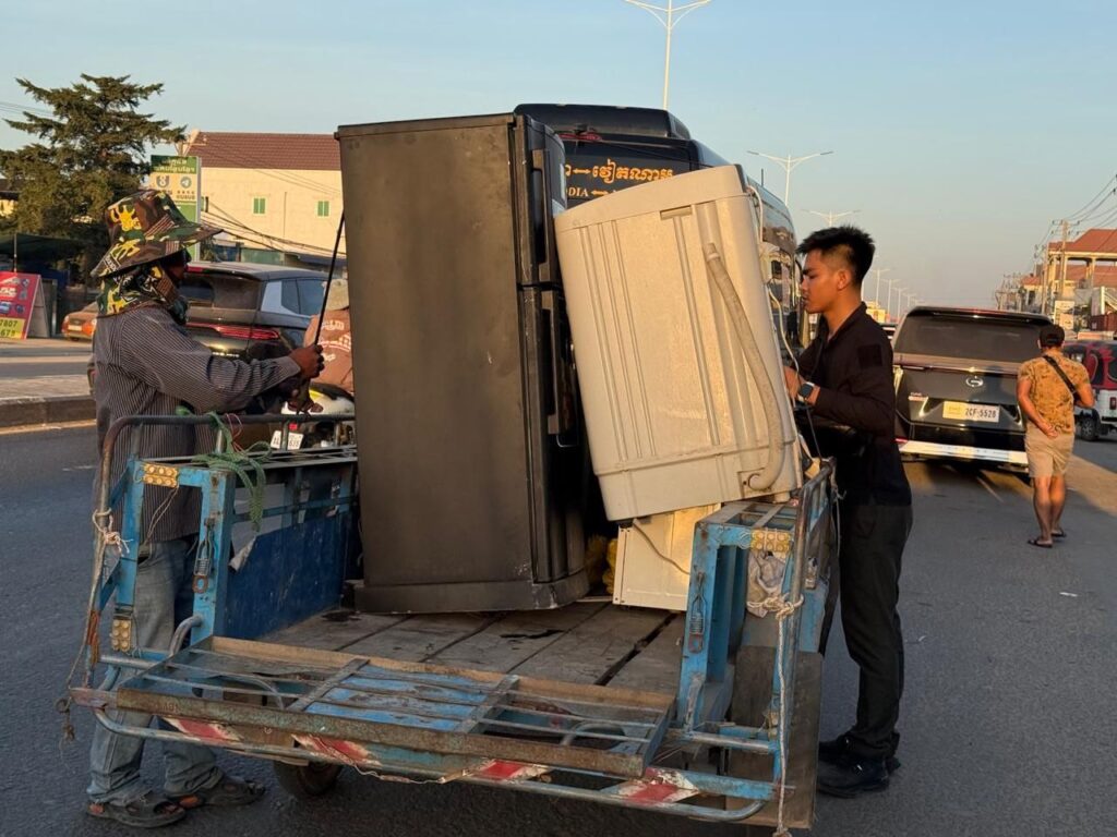 Two men load a washing machine and refridgerator into a packed motorbike trailer parked along National Road 1 in Cambodia's Bavet city on February 1, 2026. (Mech Dara/Mekong Independent)